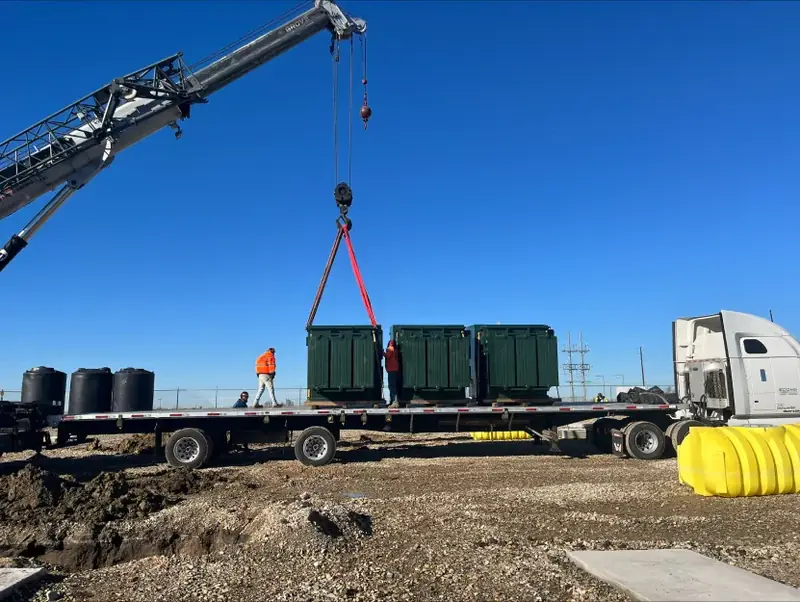 Crane unloading green equipment boxes from a flatbed trailer on a job site