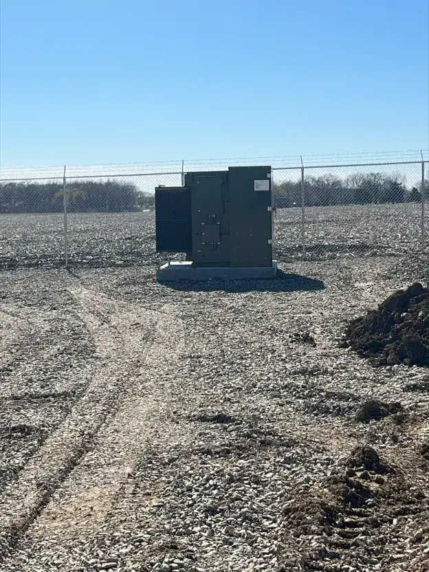 Green utility cabinet on a gravel lot by a chain-link fence