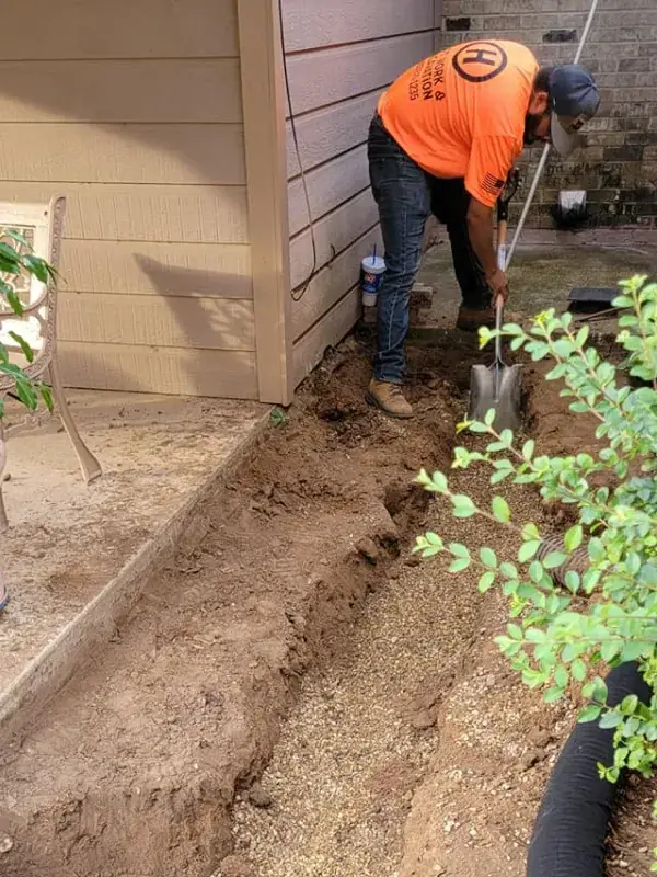 Crew member hand-digging a trench along a house, with black drain pipe at the bottom
