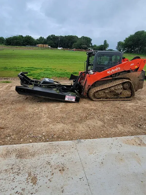 Track loader fitted with a mulching/brush-cutter head, parked beside a concrete slab